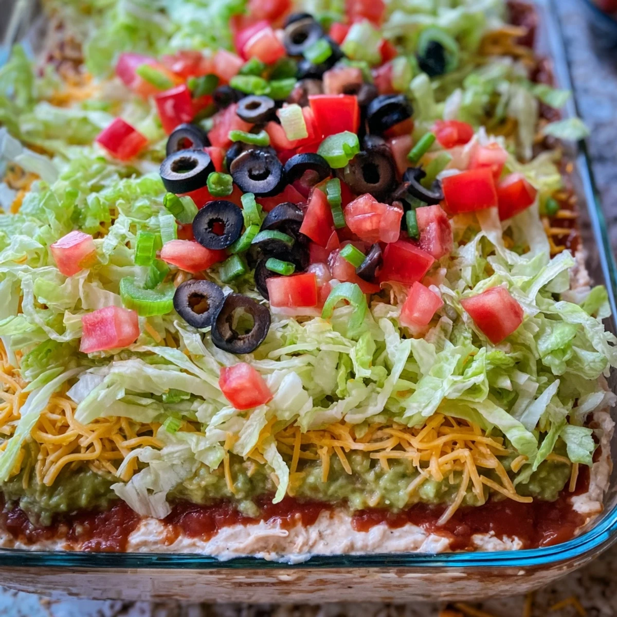 Close-up of a Super Bowl Seven Layer Taco Dip serving, with fresh lettuce, olives, and tomatoes topped on a cheesy layer.