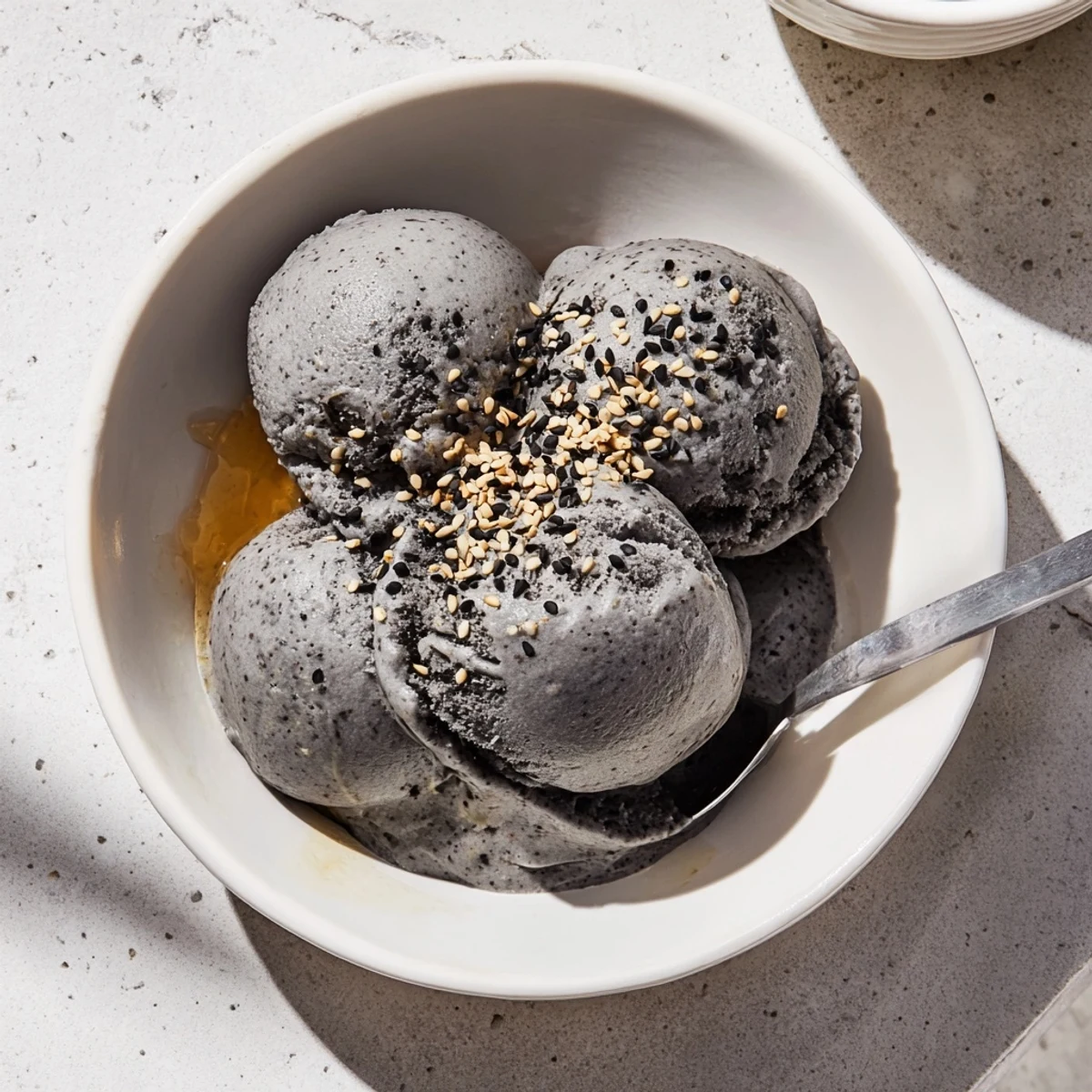 A scoop of homemade Toasted Black Sesame Ice Cream in a ceramic bowl, garnished with extra sesame seeds and a honey drizzle.