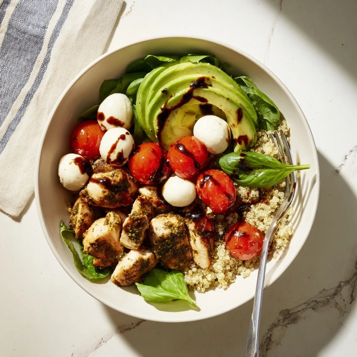 Bright grilled pesto chicken slices rest atop fluffy quinoa, next to halved cherry tomatoes, fresh mozzarella balls, and sliced avocado with a balsamic drizzle in a grain bowl.