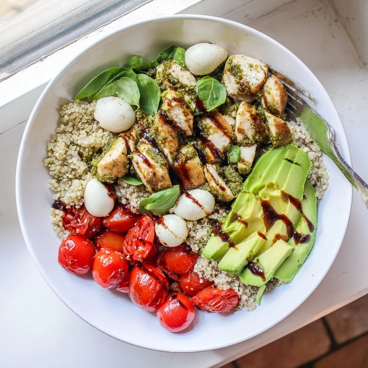 Close-up of a hearty Pesto Chicken Caprese Grain Bowl shows juicy pesto-marinated chicken, fresh mozzarella, avocado slices, and cherry tomatoes on grains with balsamic glaze and basil garnish.