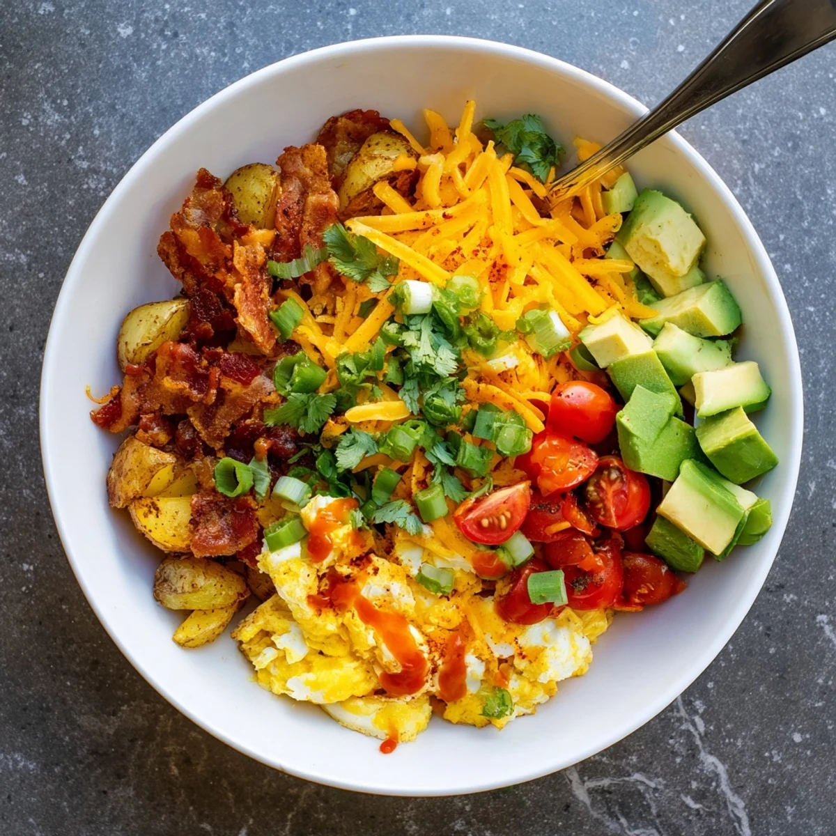 A close-up of a Loaded Breakfast Bowl with fluffy scrambled eggs, crispy roasted potatoes, and crumbled bacon topped with melted cheese.