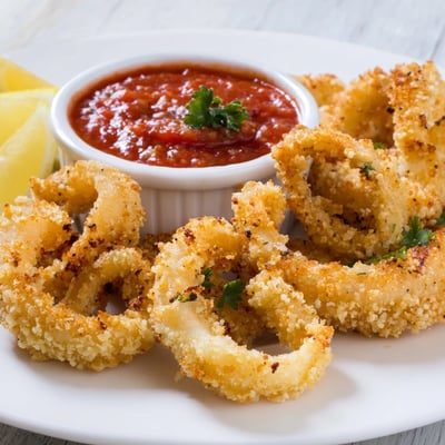 Freshly fried calamari with a golden crunch sits beside zesty marinara dipping sauce, garnished with fresh parsley and lemon for a bright Italian appetizer.  